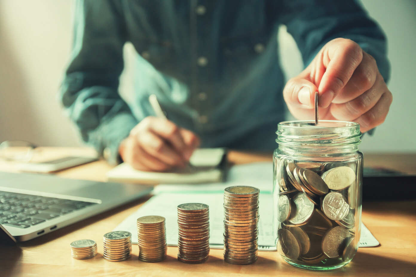 Imagem de um homem depositando uma moeda em um pote com mais moedas, ao lado dele tem mais pilhas de moedas